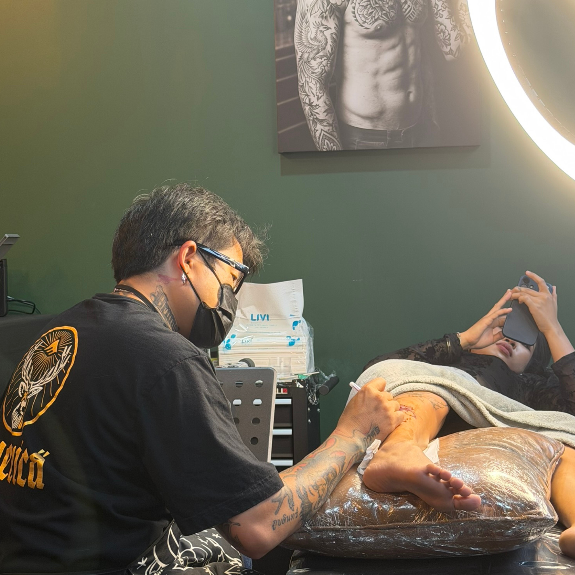 Tattoo artist working on a client's leg under a ring light in the studio